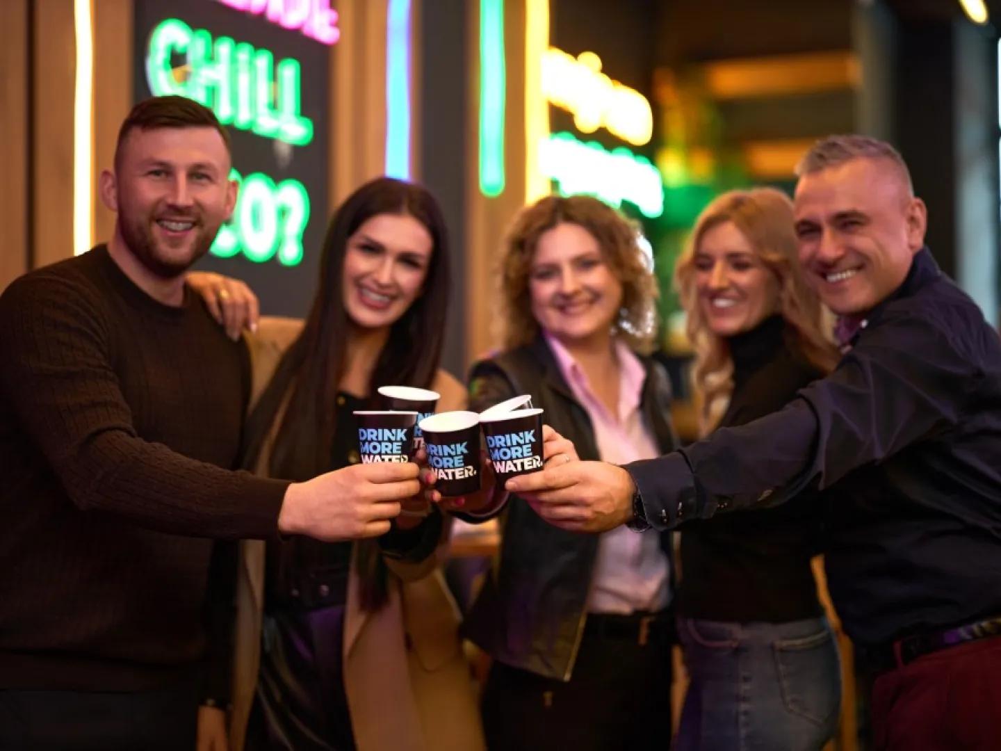 dwóch mężczyzn i dwie kobiety stukają się papierowymi kubeczkami z napisem "drink more water"