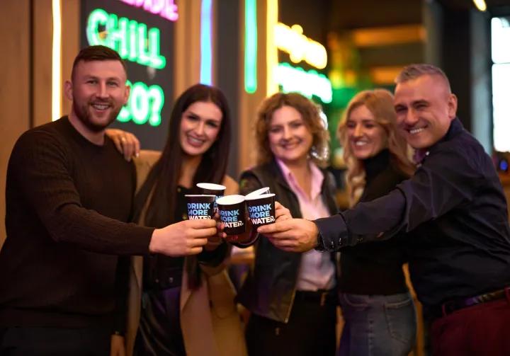 dwóch mężczyzn i dwie kobiety wznoszą toast z kubeczkami Drink more water